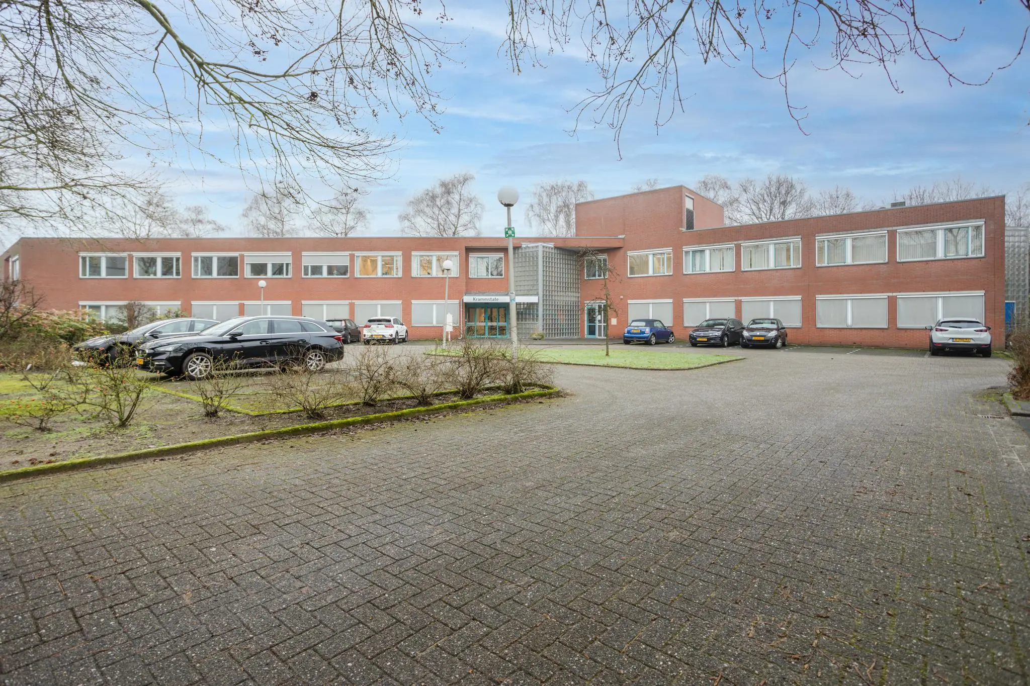 Voorgevel van een rood bakstenen kantoorgebouw aan de Christiaan Krammlaan met geparkeerde auto's op de oprijlaan.